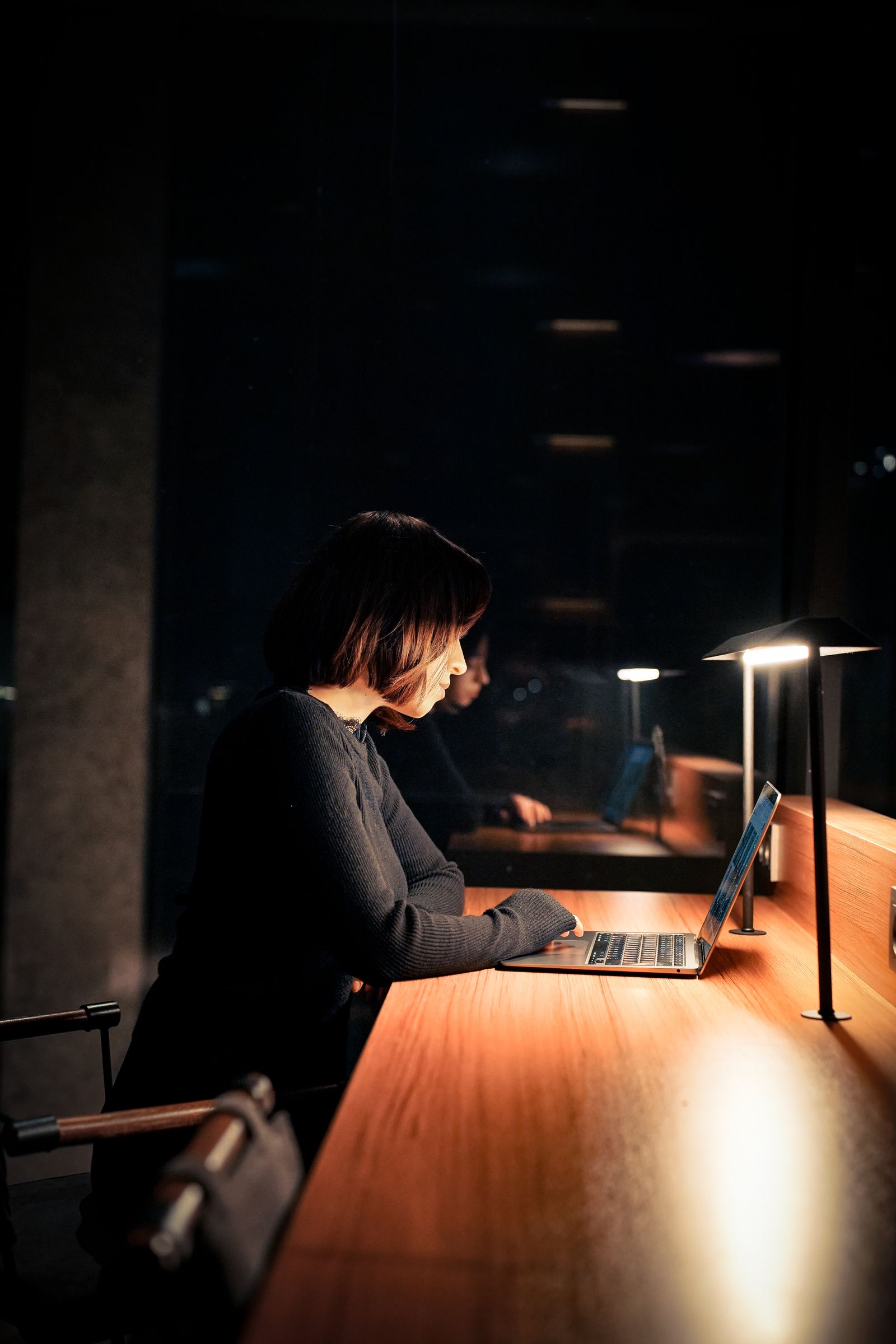 A person working at a laptop in a dim office, lit by a desk lamp.